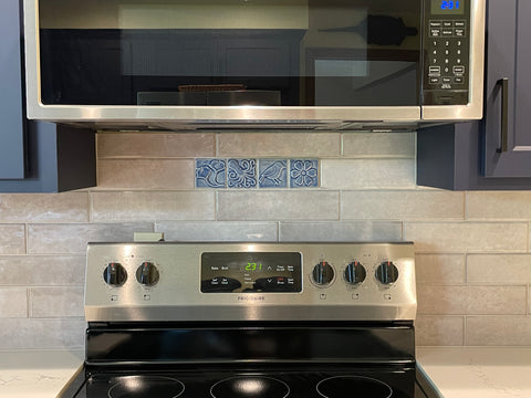 four blue handmade tiles featuring ginkgo leaves, an octopus, a chickadee and a blossom installed over a stove in a field of commercial tiles