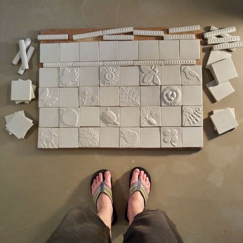 handmade tiles in white glaze laid out on the floor in front of a woman wearing rainbow flip flops