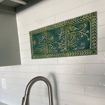 Close up of a green handmade tile mural depicting birds on a branch surrounded by a border of leaves and flowers, installed over a kitchen sink in a field of white subway tiles