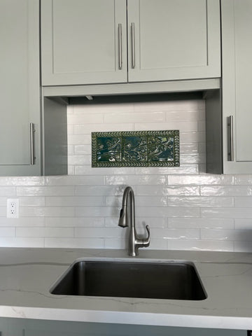 Green handmade tile mural depicting birds on a branch surrounded by a border of leaves and flowers, installed over a kitchen sink in a field of white subway tiles