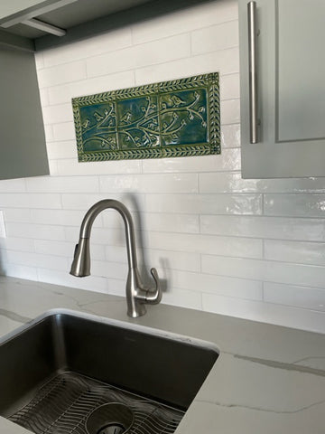 Green handmade tile mural depicting birds on a branch surrounded by a border of leaves and flowers, installed over a kitchen sink in a field of white subway tiles