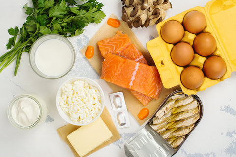 Top down view of a white table and foods high in calcium and vitamin D including salmon, eggs, canned fish, dairy, and leafy greens.