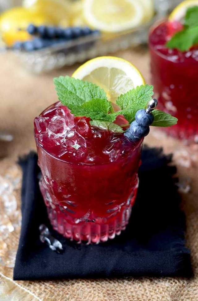 clear glass tumbler filled with dark pink liquid ice blueberries mint leaves and lemon slice