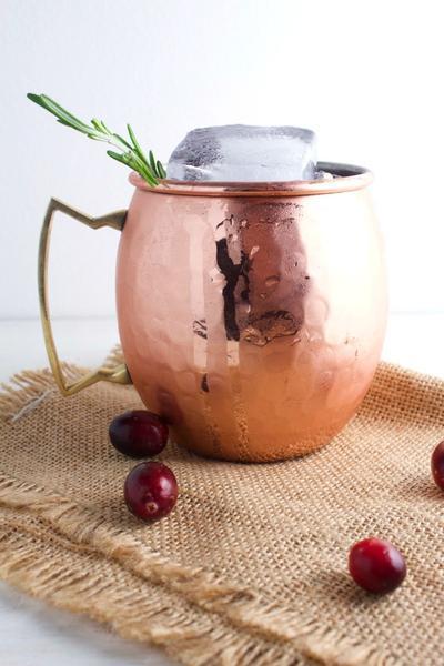 Moscow Muled copper mug filled with liquid ice sprig of herb and surrounded by cranberries side view