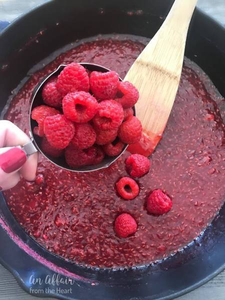 pot filled with crushed raspberries and hand holding a cup filled with whole raspberries