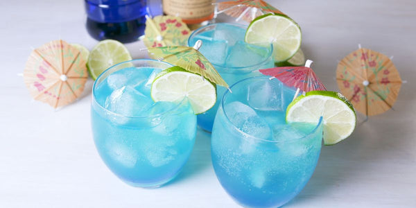 three clear glass filled with blue liquid ice cubes and sliced lime and umbrella wedged on the rim