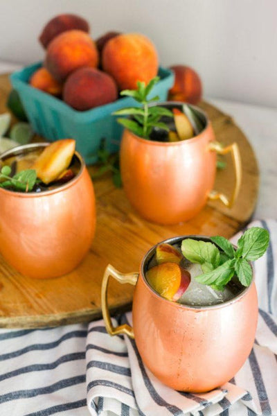 three copper mugs filled with liquid peach slices mint leaves and ice cubes