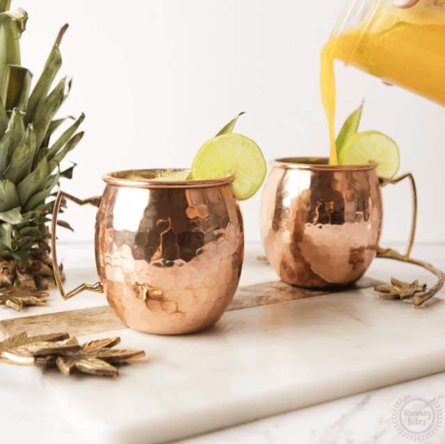 two Moscow Muled copper mugs with citrus slice on its rim and hand pouring yellow liquid into one mug