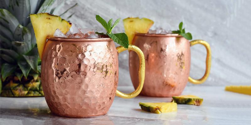 two Moscow Muled copper mugs filled with ice and pineapple and mint leaves wedged on the rim