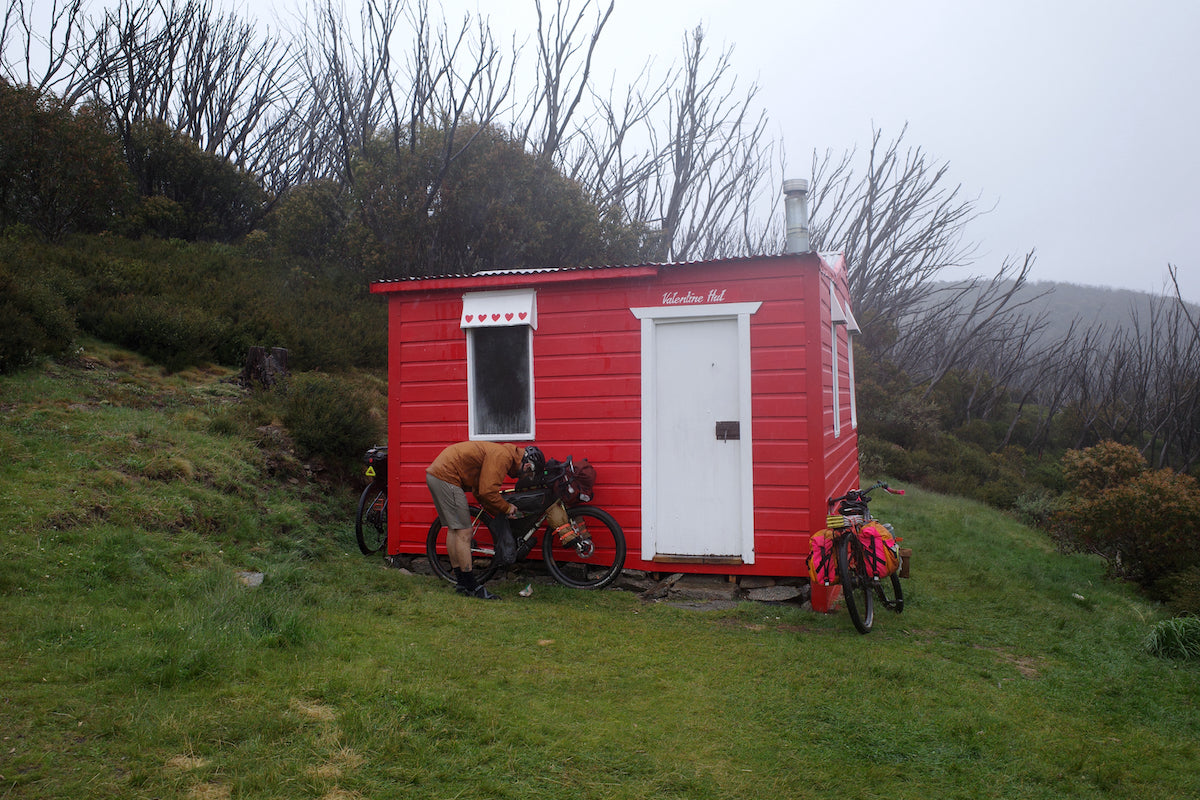 In the morning fog, a bikepacker loads his bike in preparation for the day ride. The red shed, which the bike is leaning on, contrasts nicely with the engulfing chaparral and tall grass.