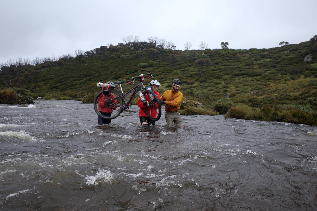 Three bikepackers fjord across a river while carrying a mountain bike above their shoulders. The rushing water and dark skies show that the weather is present and mother nature is alive.