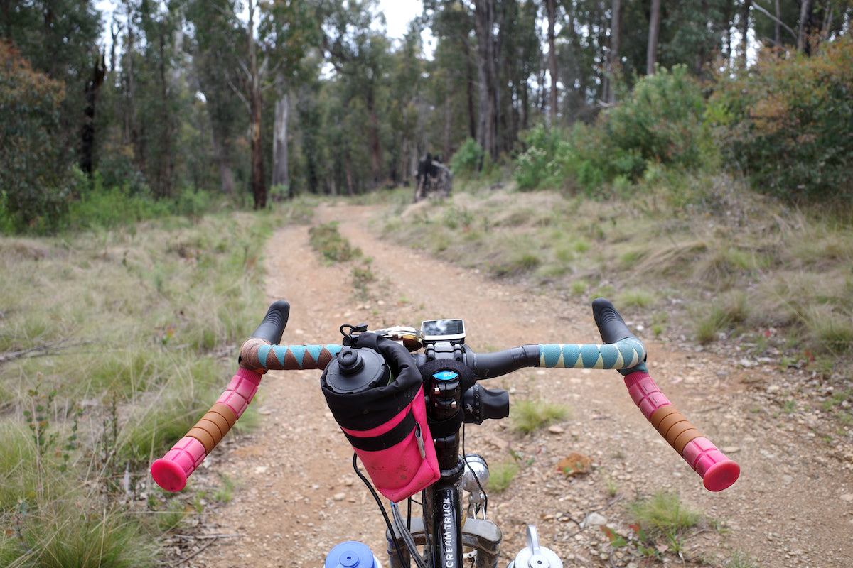 Robbie is giving us a preview of their handlebar setup. You can see their colorful bar tape, Highlighter Pick Co Pilot by Road Runner Bags, and a Wahoo mounted to the stem. The distance shows a eucalyptus forest native to the Australian Outback.