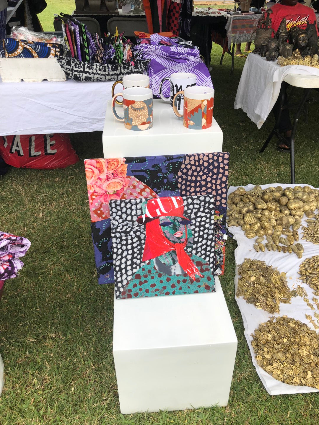 Outdoor market display with African prints, designed mugs, sculptures, and a collage art piece with a stylish woman.