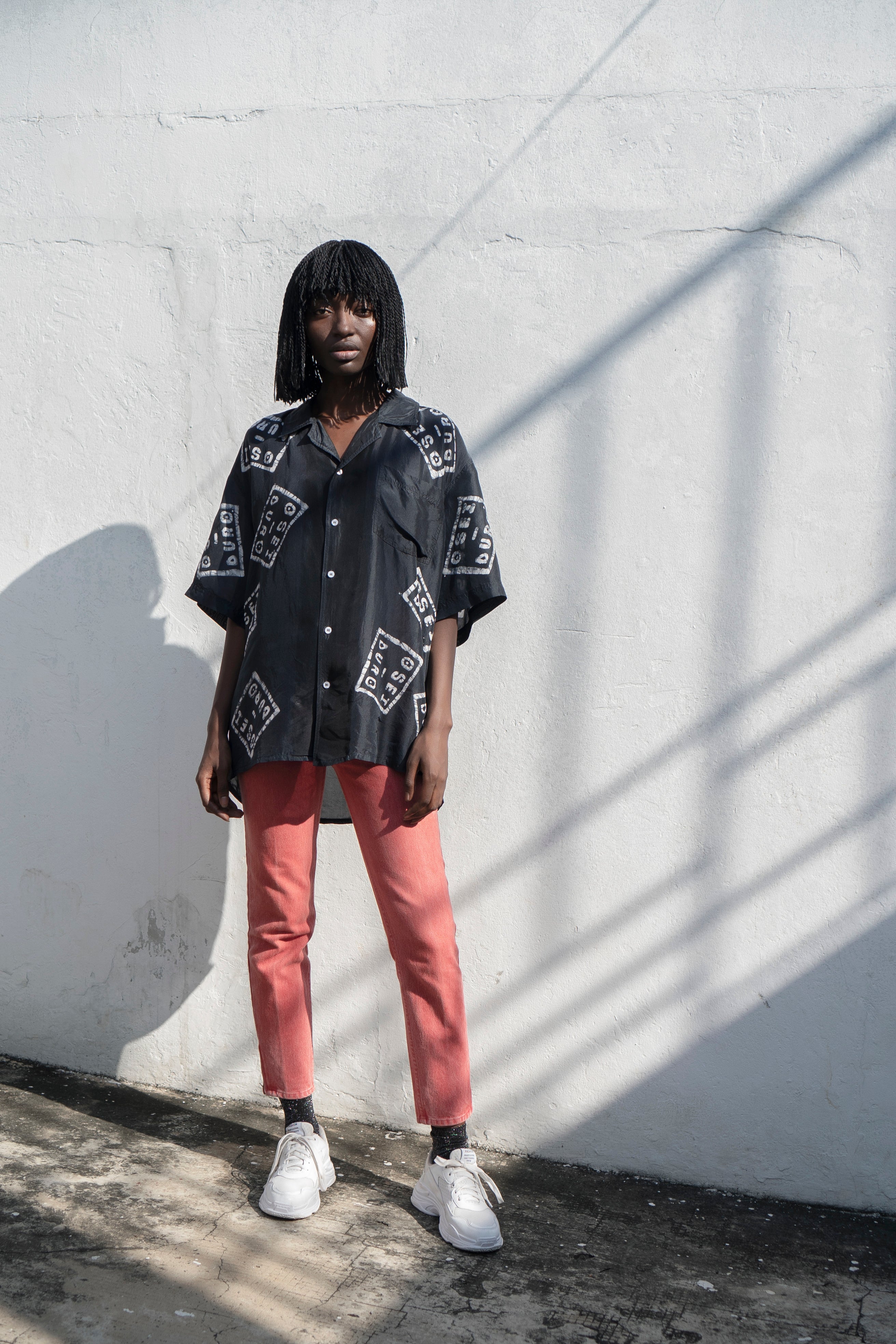 Woman in black shirt with white Osei-Duro graphic and red pants against white wall with shadows.