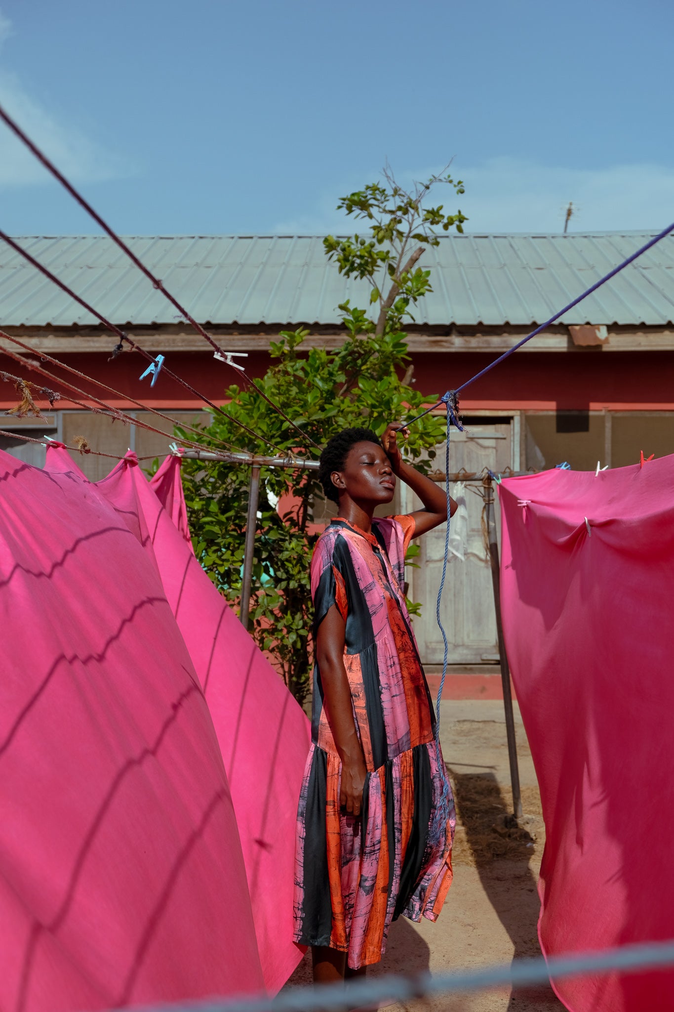 Person in a stunning batik dress, standing between pink fabrics on a clothesline, one arm raised.