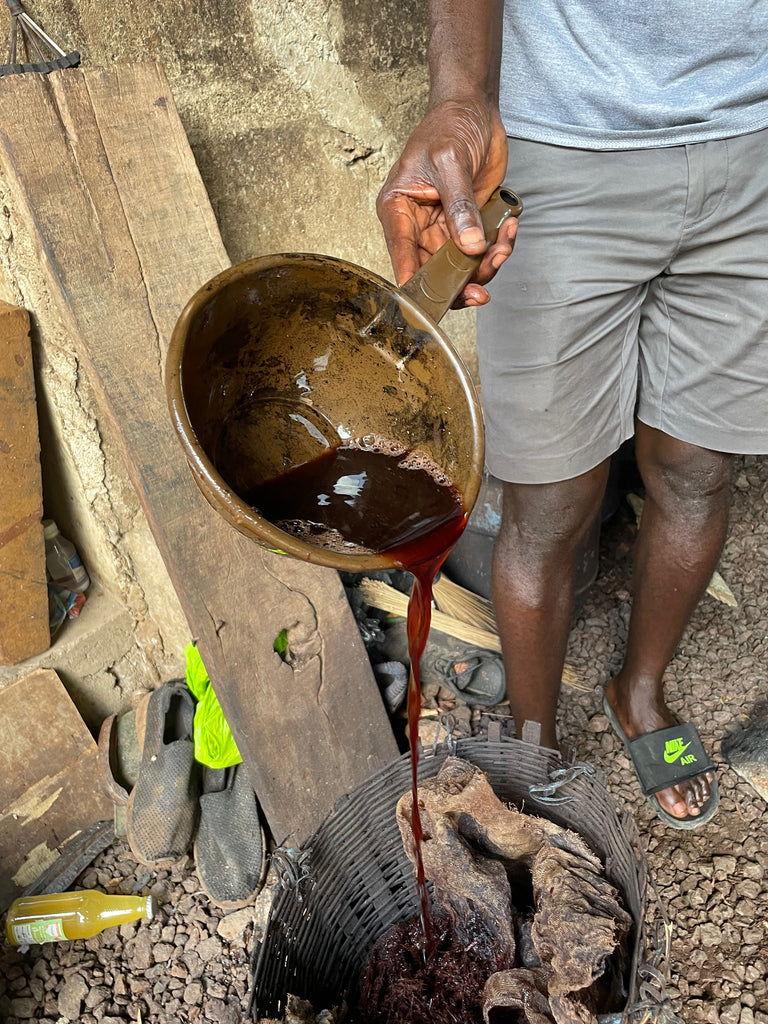 Artisan pours dark dye into basket with fabric, traditional dyeing process outdoors.