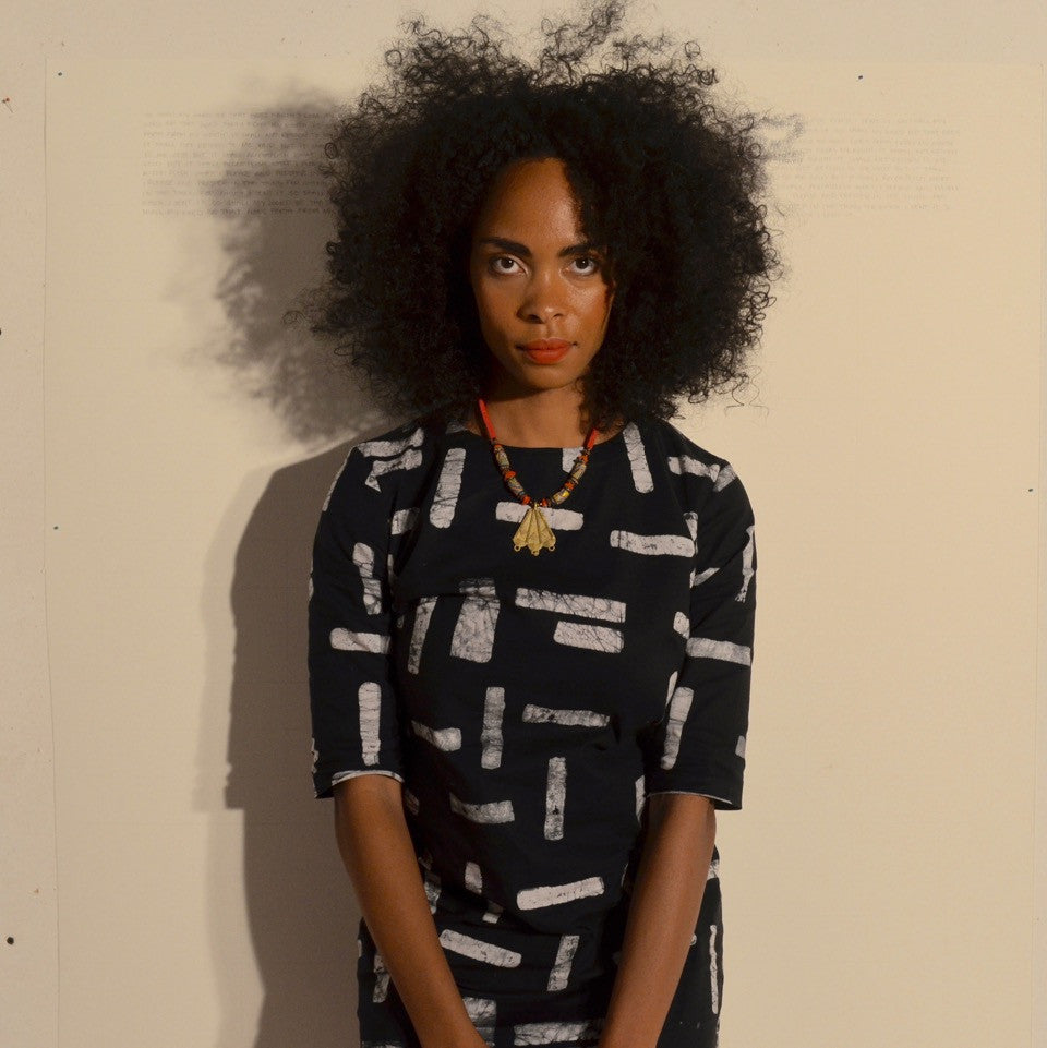 Individual with curly hair in a dark dress with white geometric patterns, standing against a light-colored wall.
