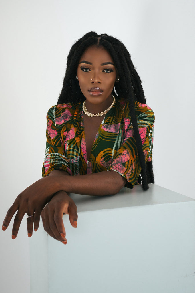 Model seated behind a white surface, wearing the Letsa Dress in Waters with dreadlocks and a gold necklace.