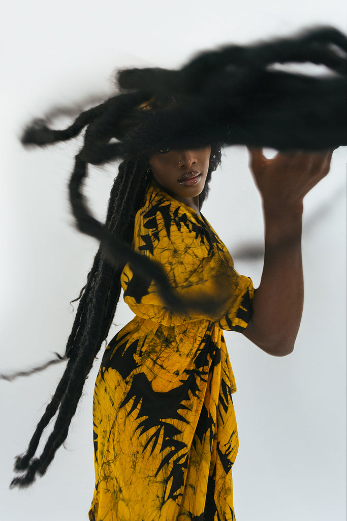 Model lifts dreadlocks, showcasing a yellow dress with black leaf patterns against a plain backdrop.