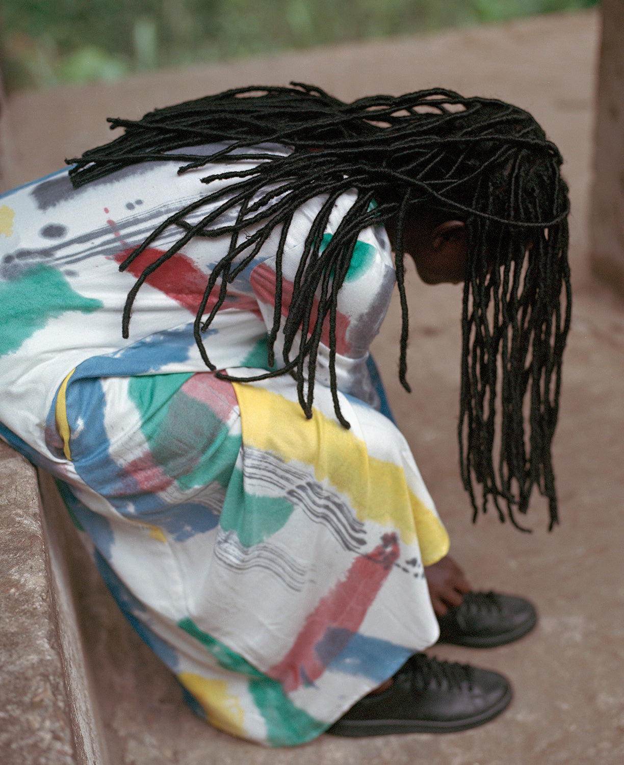 Individual in a colorful, paint-splashed Osei-Duro garment sitting on a concrete surface with head bent down tying their shoe.