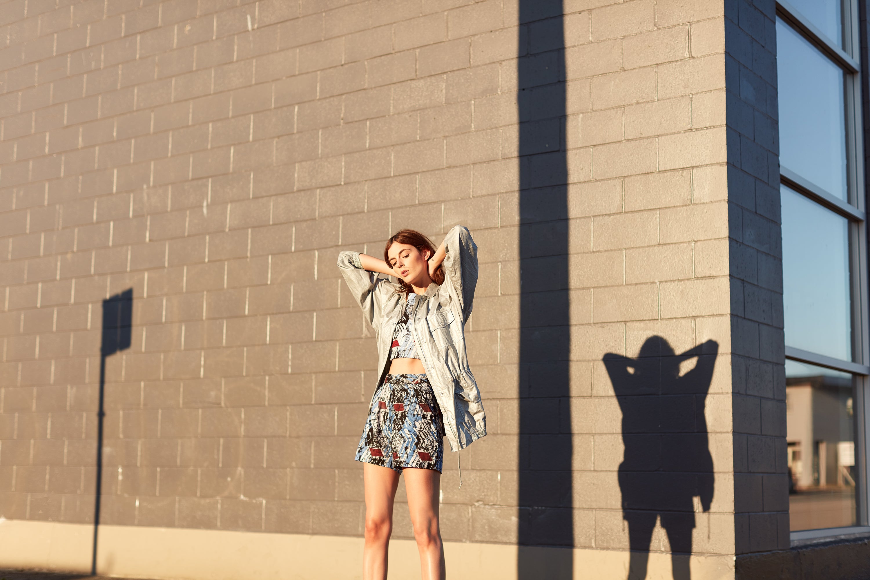 Individual standing outdoors against a plain wall with arms raised, casting a shadow on the wall, wearing an Osei-Duro skirt.