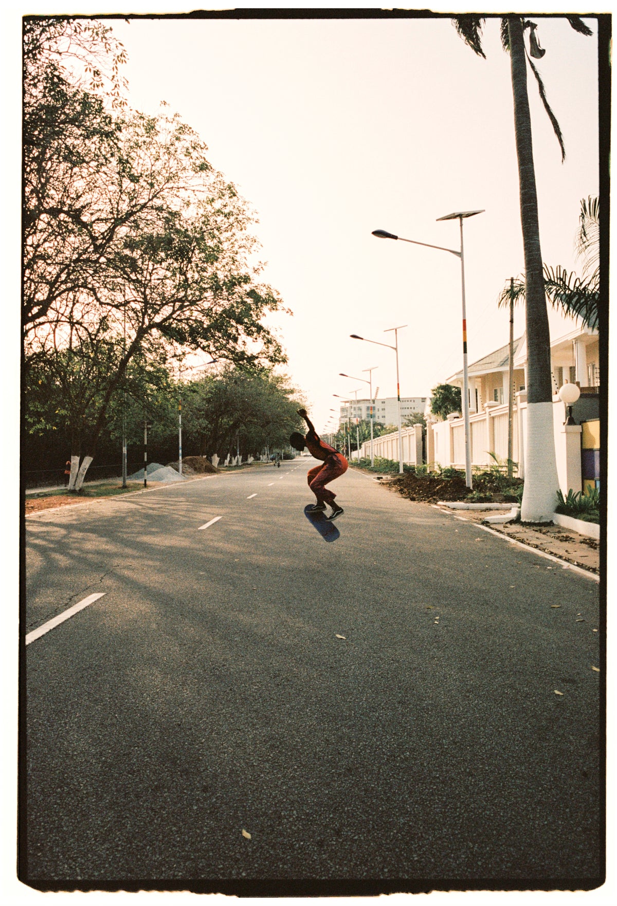 Skateboarder in Osei-Duro outfit executing a trick on a tree-lined street with clear skies.