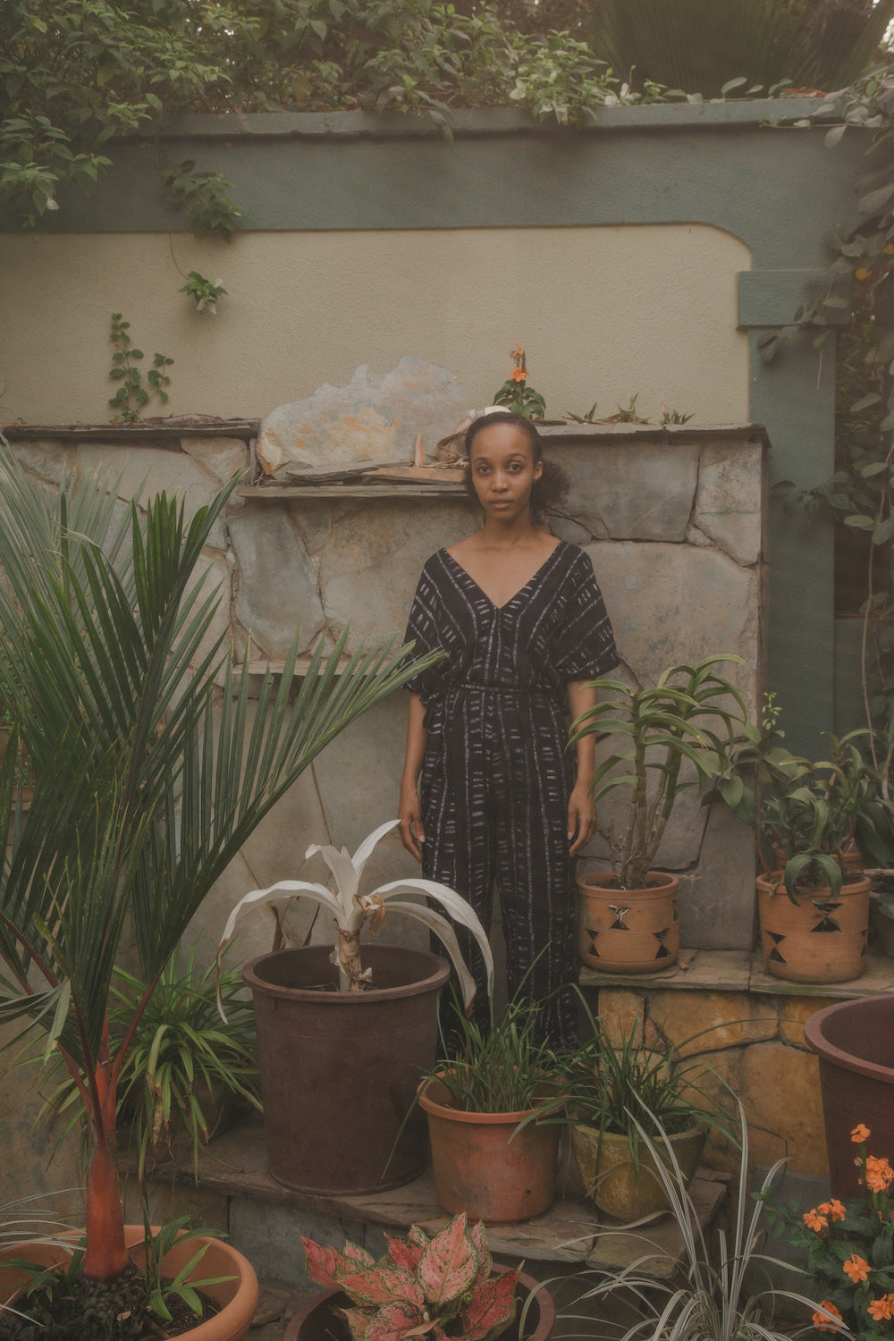 Woman stands amidst potted plants, wearing a black and white batik print jumpsuit, in a lush garden setting.