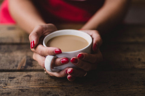 A woman holds onto a white cup with two hands, resting them on a wood table. She is wearing pink fingernail polish. The cup contains a milky coffee drink.