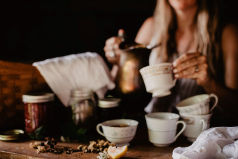 Photo of a woman pouring tea from a metal carafe into a glass tea cup. There are tea cups scattered all over a table. There is loose leaf tea and lemon wedges also on the table.
