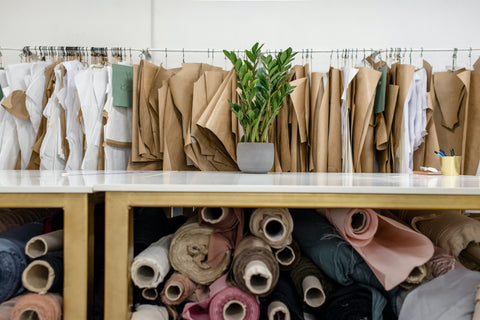 Rolls of different colored fabrics sit in a wooden shelf. Beige and white material hangs on a rack in the background. A green plant sits on a marble table.