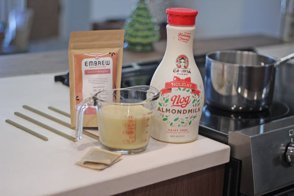 A measuring cup full of Califia Almondmilk Nog, a carton of eggnog, two tea bags, and a pouch of Embrew tea sit on a counter. 