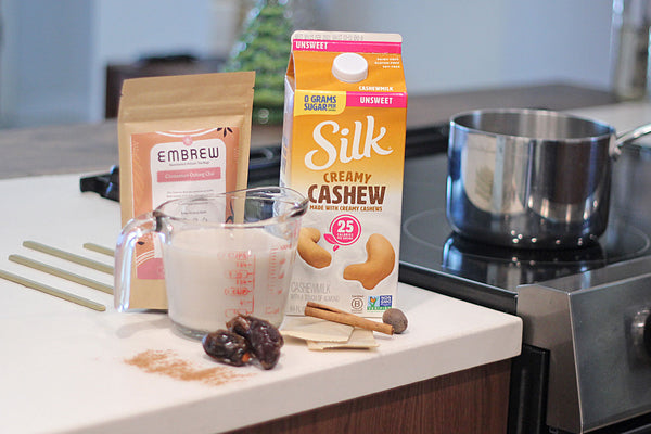 A measuring cup full of Silk Cashew milk, a carton of milk, two tea bags, and a pouch of Embrew tea sit on a counter by the stove. 