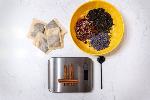 A food scale weighs some cinnamon sticks beside a teaspoon, a bowl of tea leaves, and a handful of tea bags. 