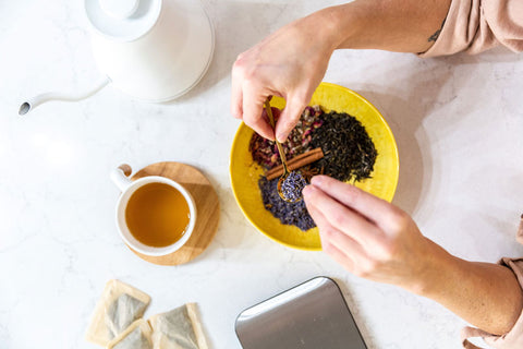 A person holds a spoonful of tea leaves over a countertop which also displays some tea bags, a mug, and electric tea kettle