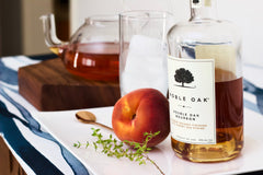 A bottle of liquor beside a ripe peach, a tea kettle, and a handful of herbs. 