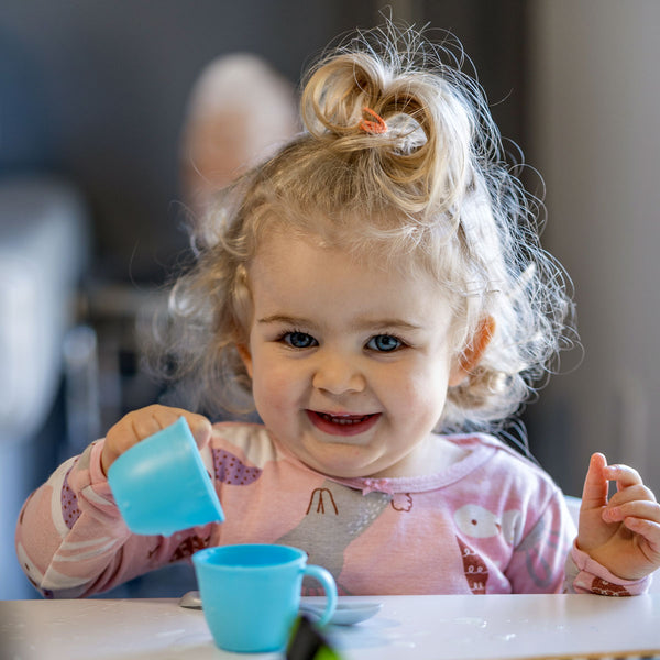 A toddler girl sits in a highchair playing with two small blue teacups. 