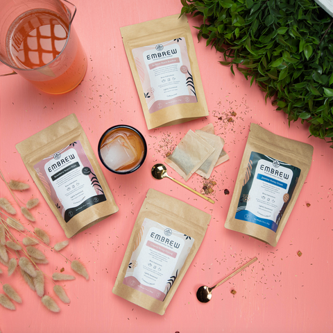 Four cold brew tea blends sit on top of a peach pink background beside teaspoons, tea bags, a pitcher, and a glass.