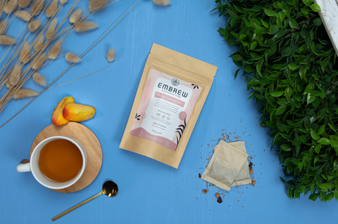 A pouch of Embrew's Summer Peach Rooibos sits on a blue bakcground with two tea bags, a mug, some peaches, and a teaspoon. 