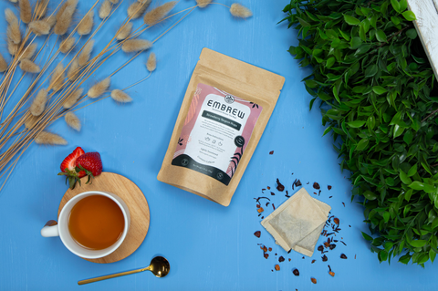 A pouch of Embrew's Strawberry Yaupon Hops Sweetened Tea on a blue background beside two tea bags, a teaspoon, and a tea cup. 