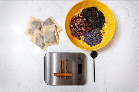 A handful of Embrew tea bags next to a scale and a bowl of loose leaf tea. 