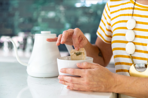 A woman sits at a counter and steeps tea with a tea bag. 