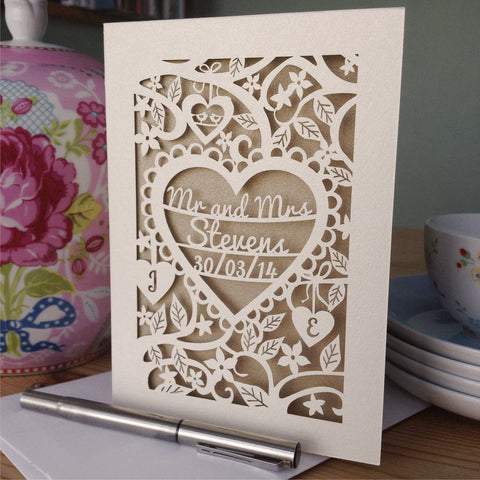 A Personalised Papercut wedding Card featuring the words "Mr and Mrs Stevens" and the date inside a heart, surrounded by flowers and leaves. There are two hearts underneath with an initial in each heart.