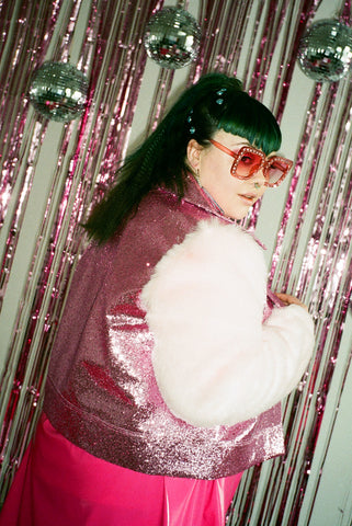 Plus size beautiful street cast model wears Elton John style pink sunglasses and glitter biker jacket with faux fur sleeves and a pink pvc skirt