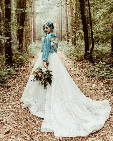 beautiful bride with blue hair stands in a woodland clearing in her wedding dress with a blue sequin biker jacket and fresh cut flowers