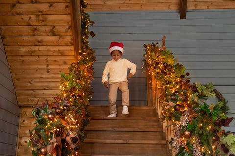 A young boy in a Santa hat walks down a staircase decorated with garland on Christmas morning