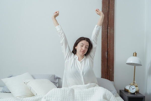 A woman that’s sitting up on her bed while stretching after a good sleep.