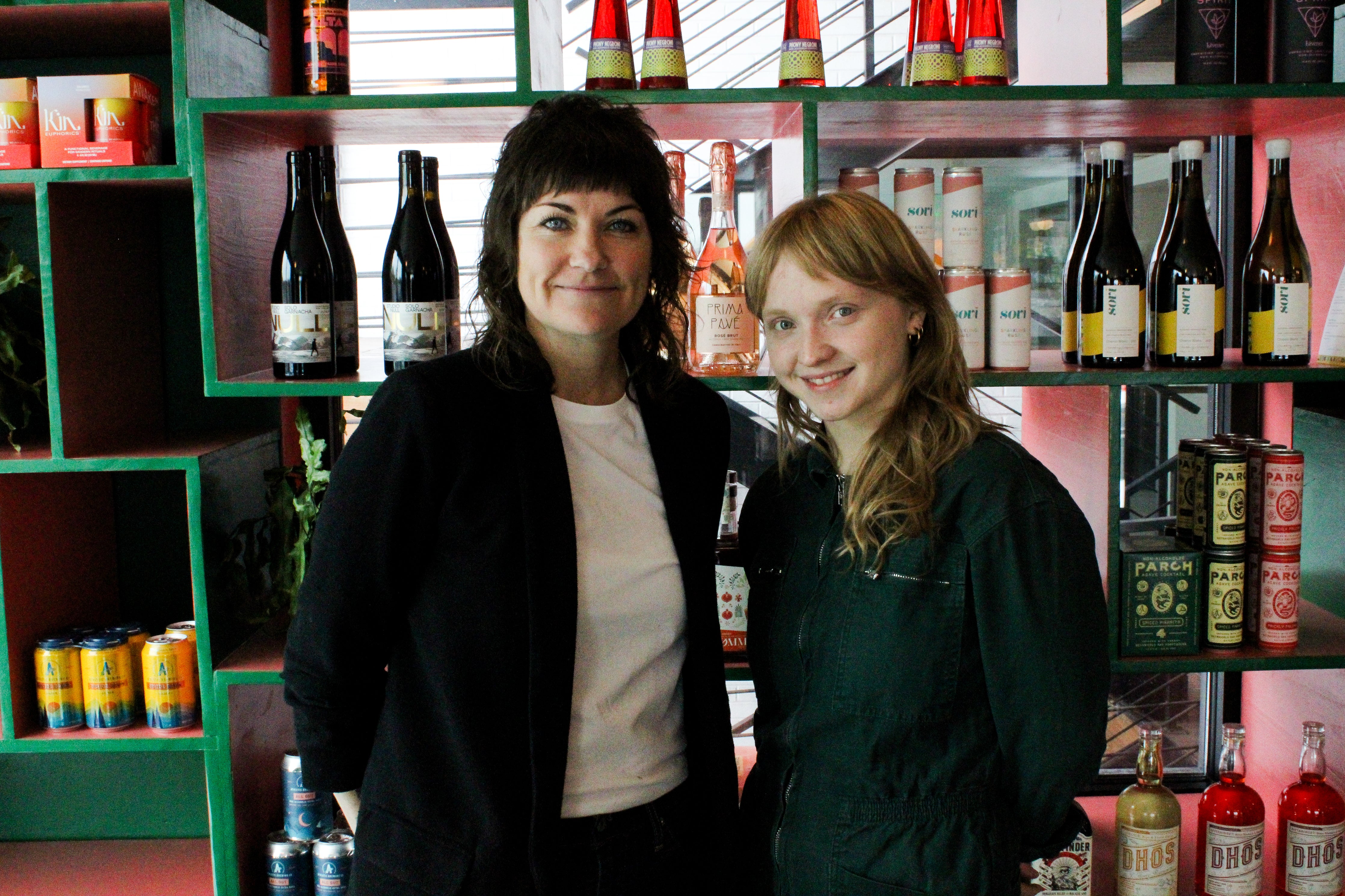 Two women stand in front of a colorful bottle display at Curiosity.