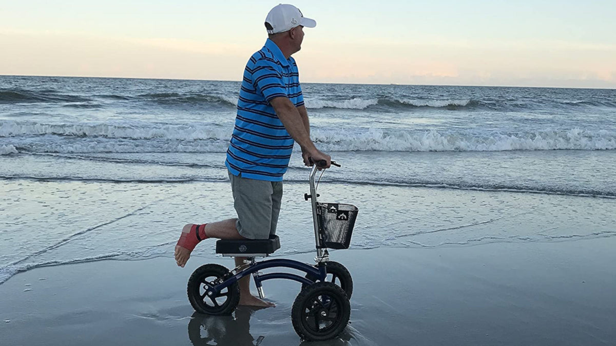 a man in a blue tshirt and shorts using a knee scooter to walk along the beach at twilight