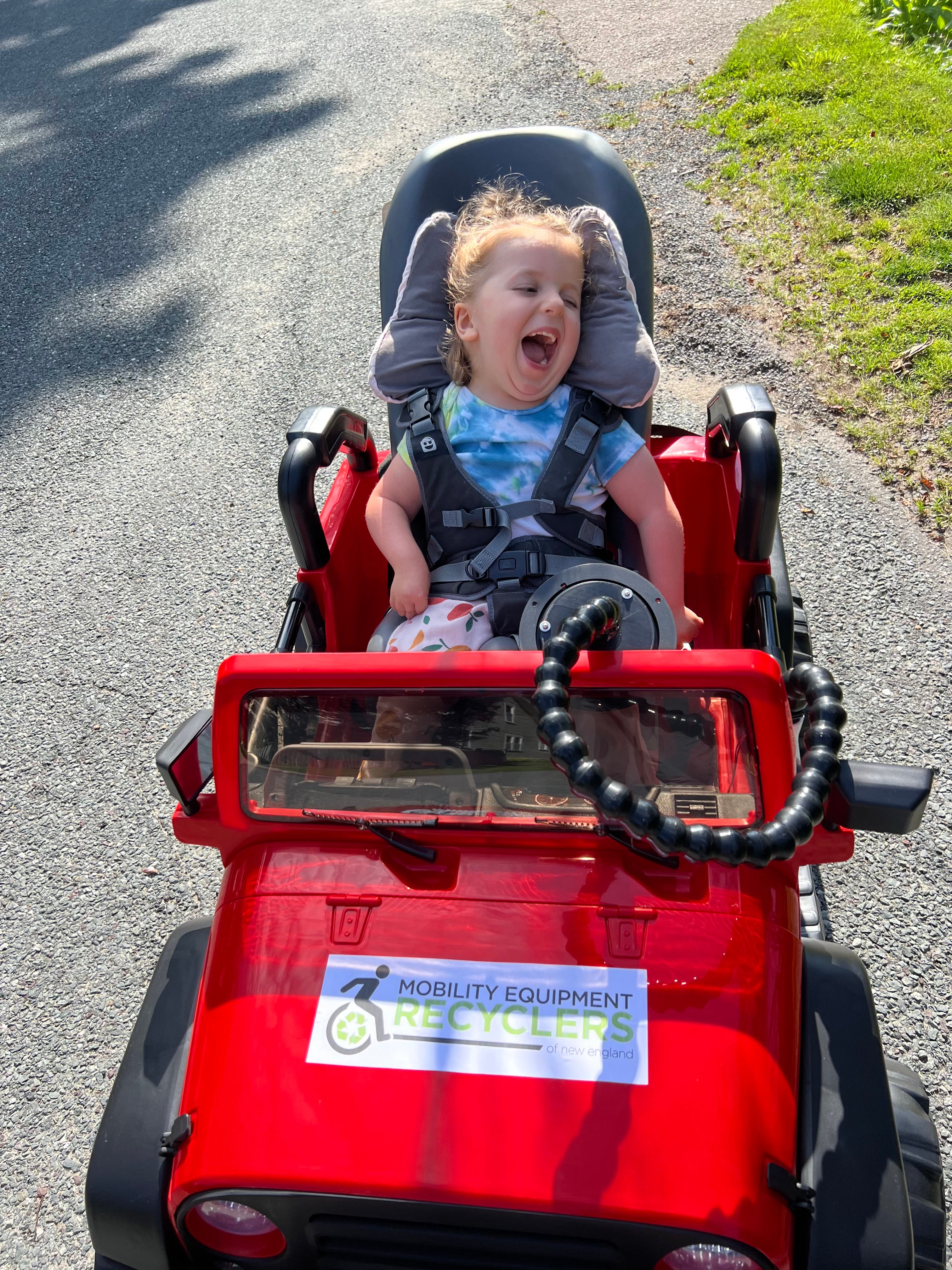 Bear, a young girl with blond hair wearing a tie-dyed tshirt, is laughing while sitting in her specially adapted red motorized Jeep