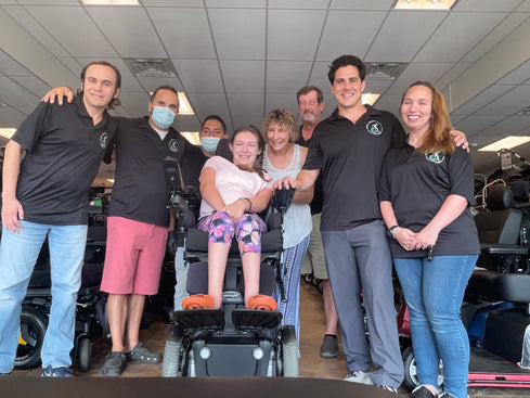 Laura, a young woman in a pink top and purple leggings, poses for a group photo in her new wheelchair, surrounded by her mother and the Mobility Equipment Recyclers staff